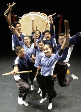 Group of students gather for photo with drum sticks in hand on stage in front of a drum